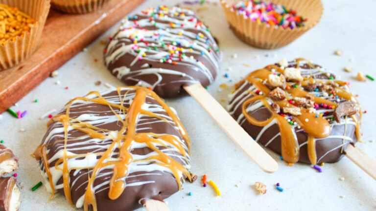 Chocolate-covered apple slices on sticks, drizzled with caramel and white chocolate, topped with colorful sprinkles and nuts.