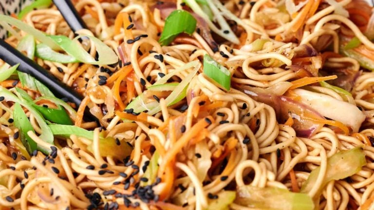 Close-up of a plate of stir-fried noodles with vegetables, garnished with green onions and black sesame seeds. Chopsticks are visible on the side.