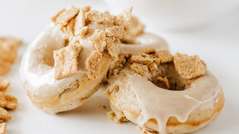 Two glazed donuts topped with crushed cereal on a white surface.