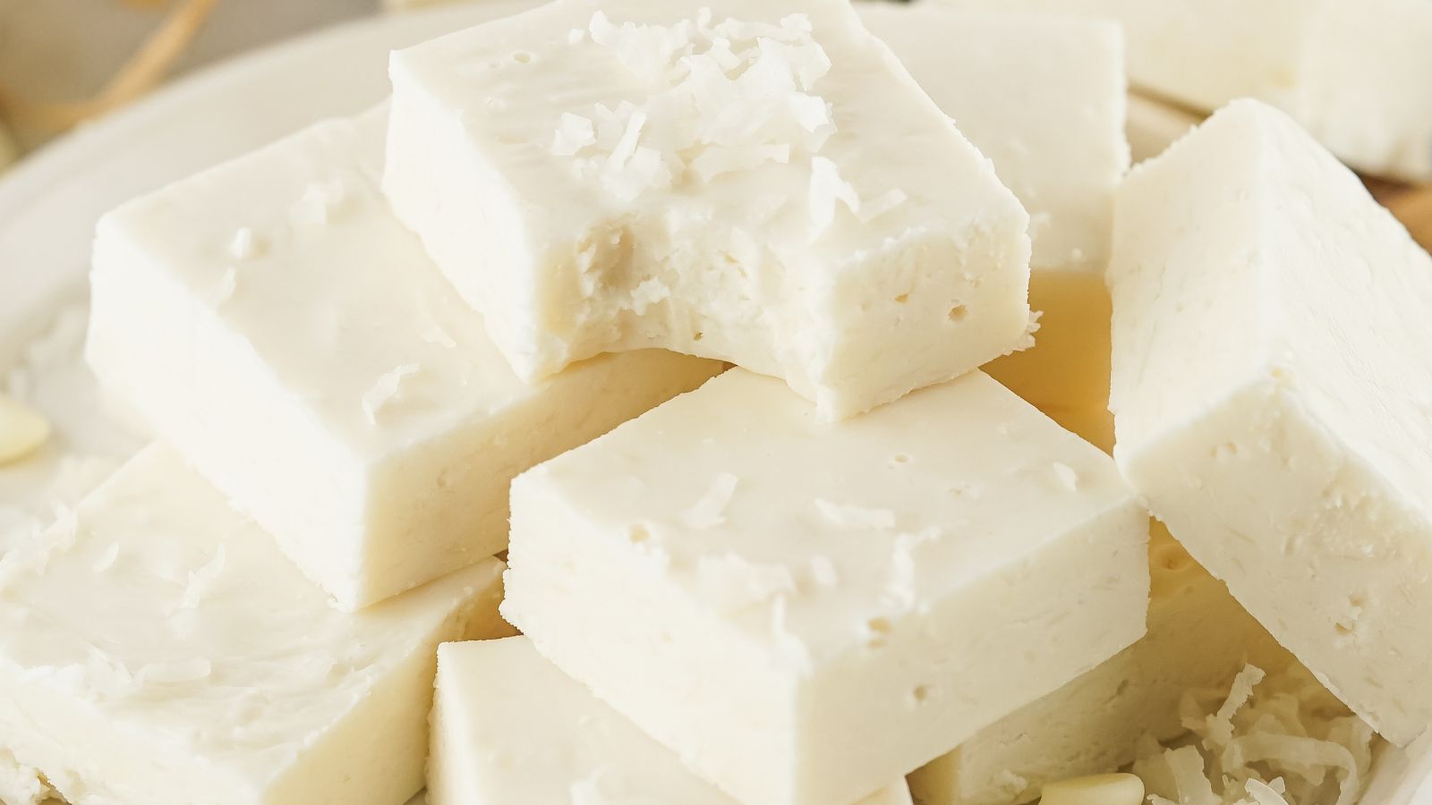 A stack of white, square-shaped fudge pieces with a creamy texture are arranged on a plate, some with coconut flakes on top.