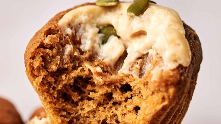 Close-up of a pumpkin muffin with a bite taken out, topped with cream cheese frosting and pumpkin seeds.