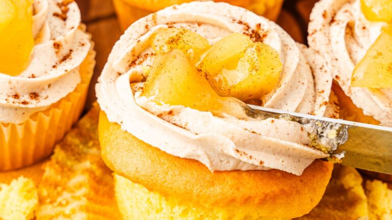 Close-up of cupcakes with swirled frosting topped with diced apples and a sprinkle of cinnamon. A knife cuts into one cupcake.
