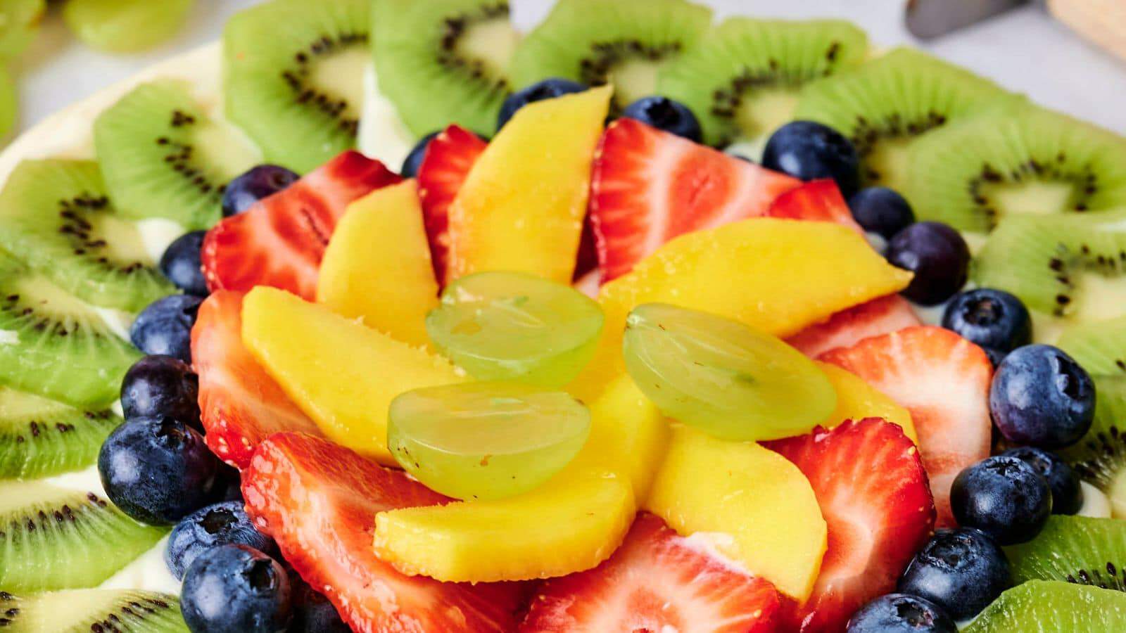 A close-up of a vibrant fruit tart topped with slices of kiwi, strawberries, mango, grapes, and blueberries arranged in a decorative pattern.