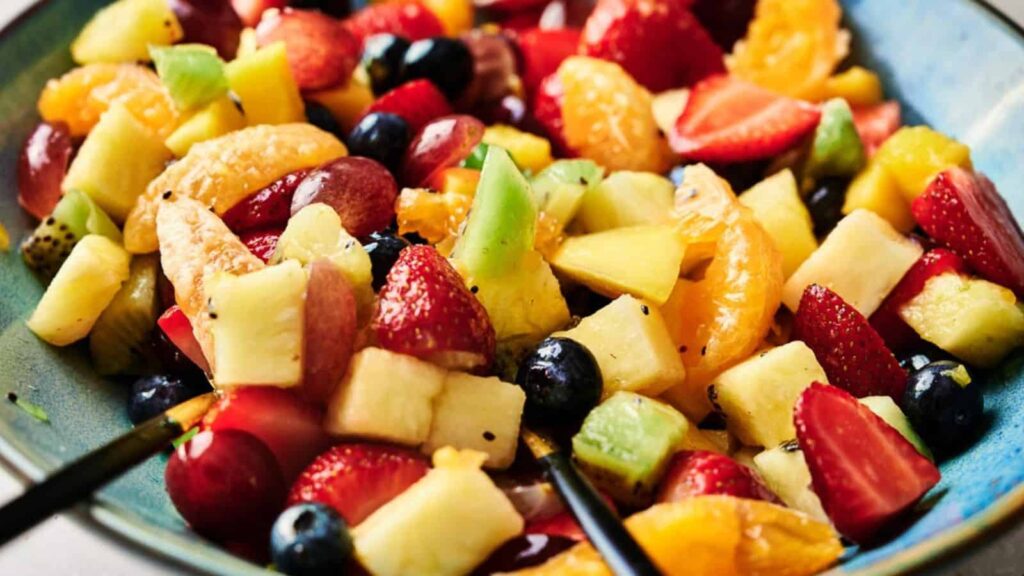A bowl of colorful fruit salad contains strawberries, blueberries, pineapple, kiwi, grapes, and orange slices. Two spoons rest inside the bowl.