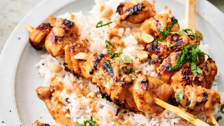 Grilled chicken skewers with peanuts and herbs sit on a bed of rice, topped with a creamy sauce.