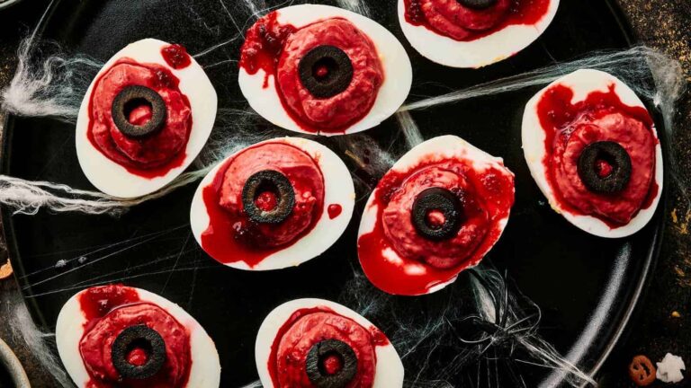 Deviled eggs styled as spooky eyes with olive slices and red sauce on a dark plate, surrounded by fake spider webs.