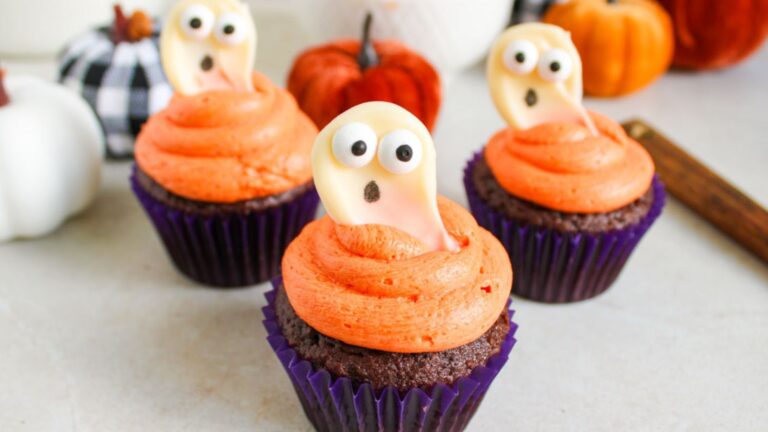 Three cupcakes with orange frosting, each topped with a small ghost decoration made of candy, set against a background featuring pumpkins and Halloween-themed items.