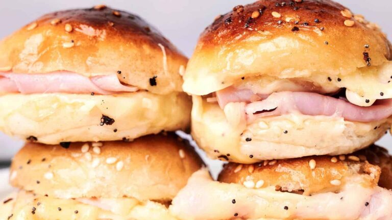 Four ham and cheese sliders with sesame seed buns stacked together.