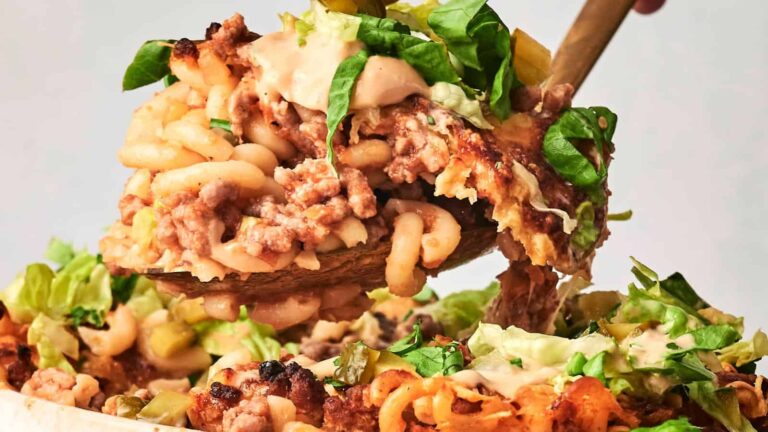 A serving spoon lifts a portion of baked pasta with ground meat, melted cheese, and fresh green lettuce garnish.