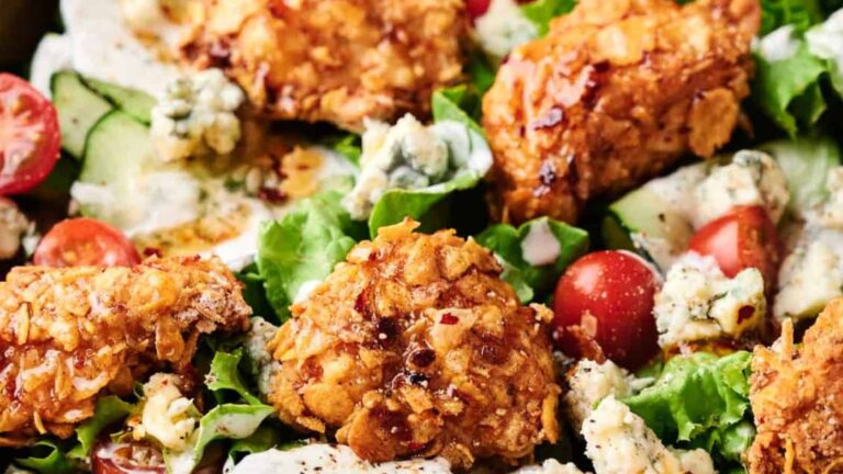 A close-up of a salad featuring crispy fried chicken pieces, cherry tomatoes, cucumber slices, blue cheese crumbles, and lettuce, all topped with a creamy dressing.