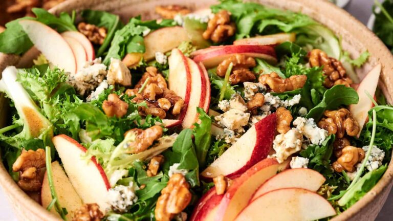 A salad with mixed greens, sliced apples, walnuts, and crumbled blue cheese in a wooden bowl.