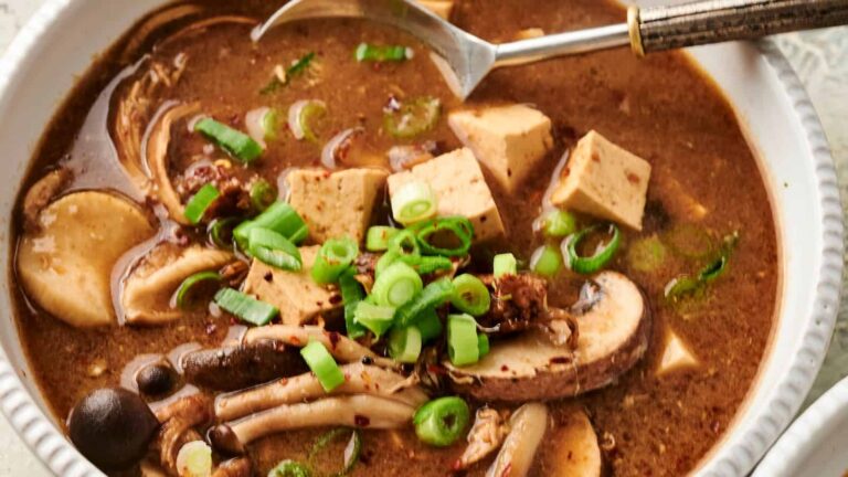A bowl of hot soup with tofu, mushrooms, and green onions, garnished with chili flakes.