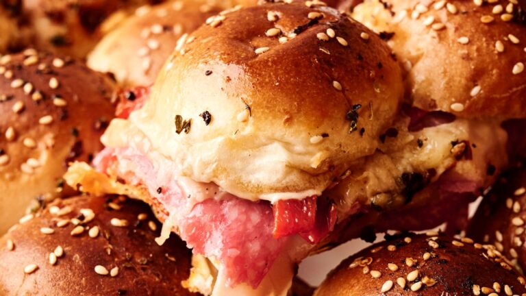 Close-up of a sandwich made with a sesame seed bun, filled with layers of meat and melted cheese, surrounded by similar sandwiches.