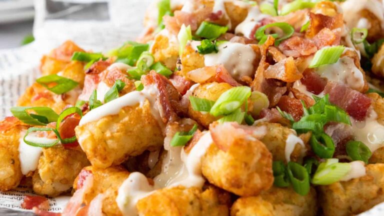 A close-up of loaded tater tots topped with melted cheese, bacon pieces, scallions, and a drizzle of creamy sauce.