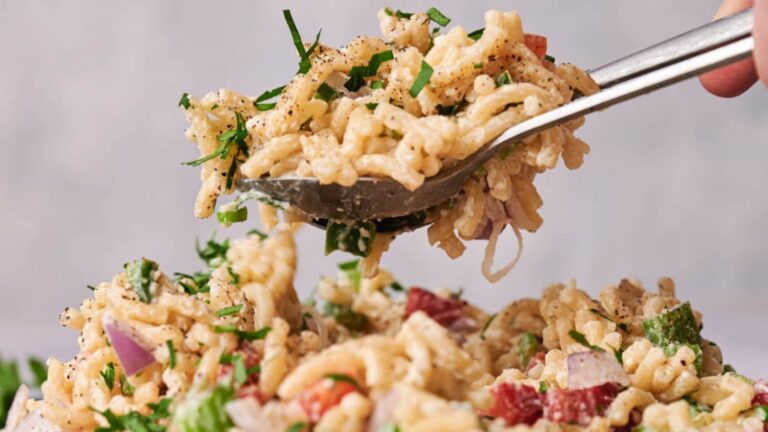 Close-up of a fork lifting a serving of creamy pasta salad with chopped vegetables and herbs.