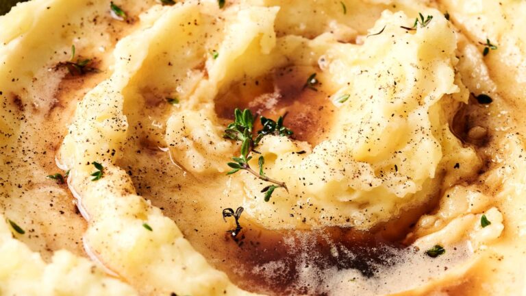 Close-up of creamy mashed potatoes with a swirl of brown gravy, garnished with black pepper and fresh thyme leaves.