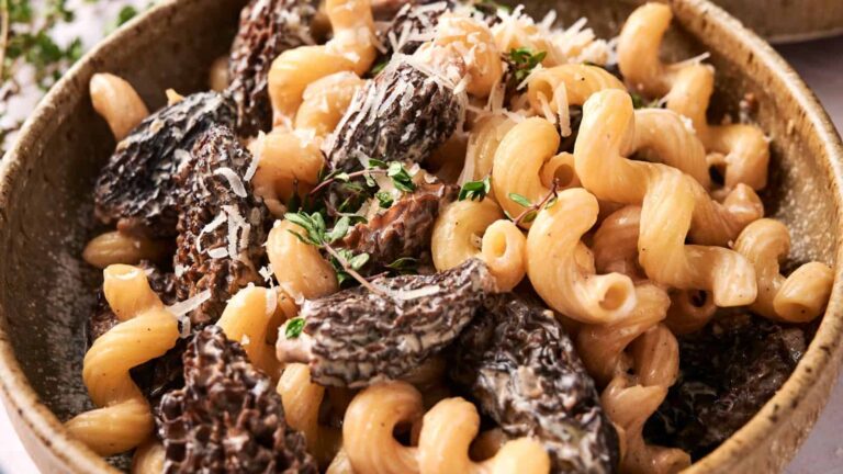 A close-up of pasta with morel mushrooms, topped with grated cheese and garnished with fresh herbs in a brown bowl.