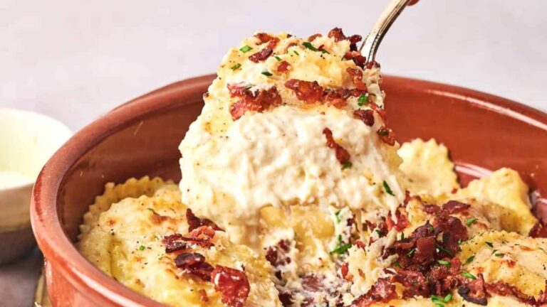 A dish of ravioli topped with creamy sauce, bacon bits, and chopped herbs, with a spoon lifting a portion.