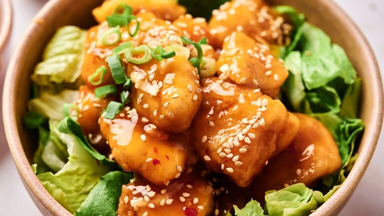 A bowl of chicken tossed in a brown sauce, topped with sesame seeds and green onions, served on a bed of lettuce.