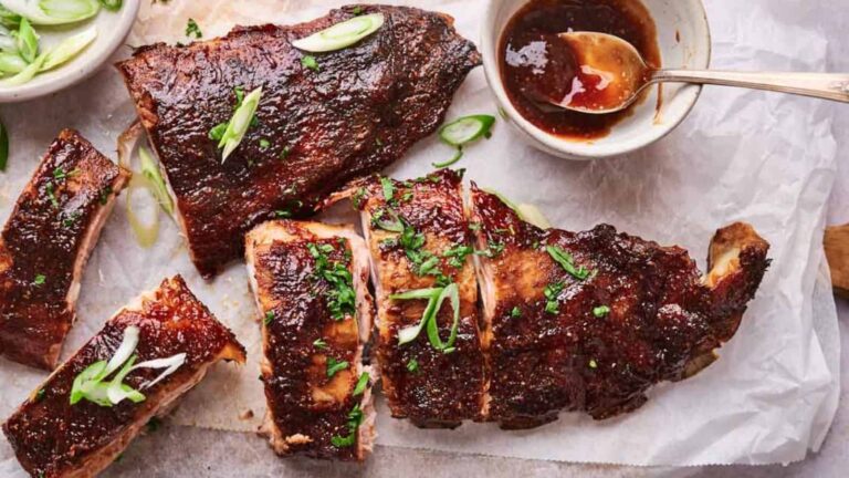 Grilled ribs with a glossy glaze are garnished with green onions and herbs, served on parchment with a bowl of sauce and a spoon.