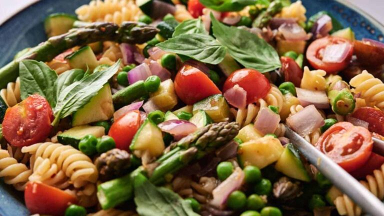 Close-up of a pasta salad featuring rotini, cherry tomatoes, asparagus, peas, red onion, zucchini, and basil leaves on a blue plate.