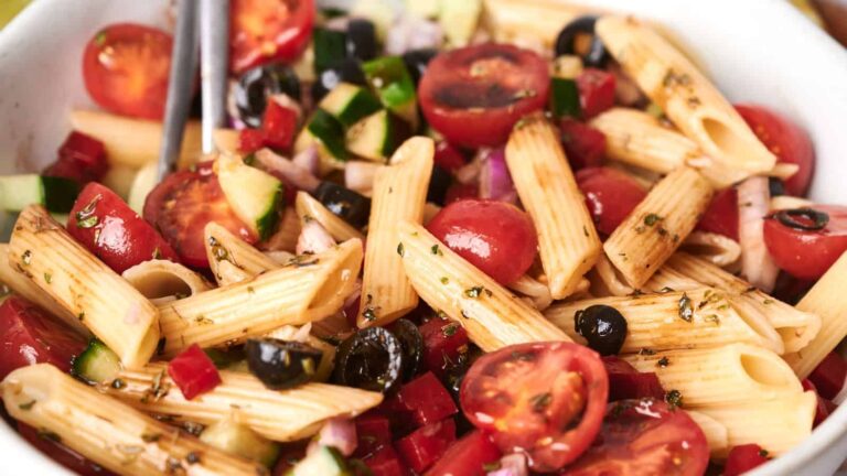A pasta salad with penne, cherry tomatoes, cucumbers, black olives, red onions, and herbs, drizzled with dressing.