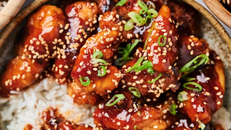 Close-up of glazed chicken pieces garnished with sesame seeds and chopped green onions, served over white rice in a bowl.
