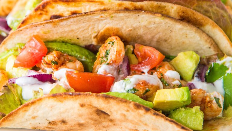 Close-up of shrimp tacos with tomato, avocado, onion, and lettuce in a toasted shell, topped with white sauce.