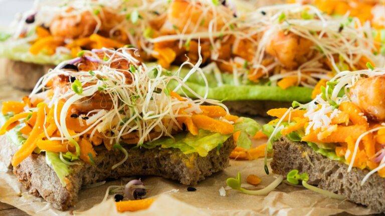Close-up of open-faced sandwiches with avocado, grilled chicken, shredded carrots, and sprouts on whole grain bread, placed on parchment paper.