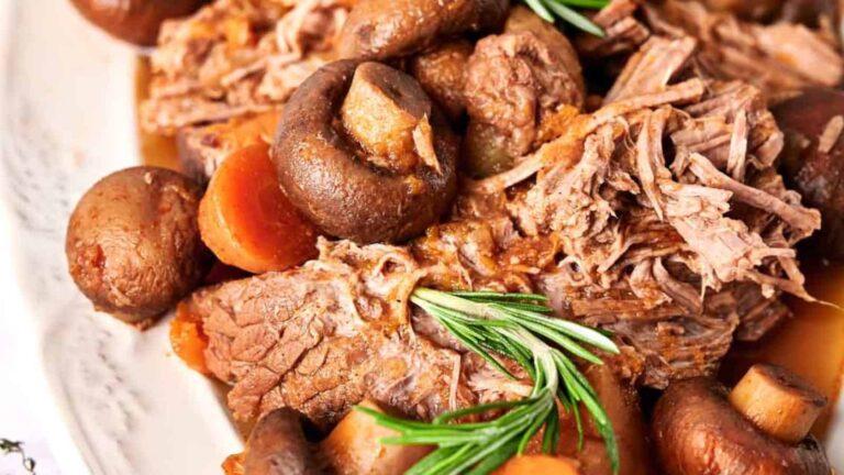 A close-up of cooked beef with mushrooms, carrots, and rosemary on a plate.
