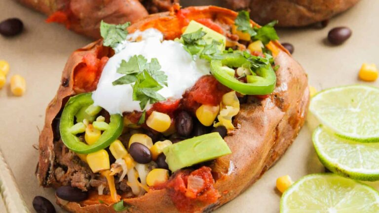 Loaded baked sweet potato topped with black beans, corn, avocado, jalapeños, sour cream, salsa, and cilantro, with lime slices on the side.