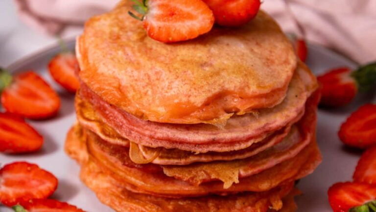 A stack of pancakes topped with sliced strawberries, surrounded by additional strawberry slices on a plate.
