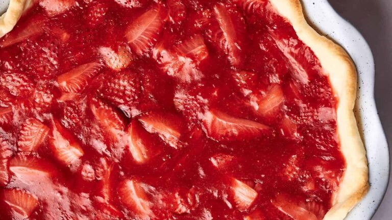 Close-up of a strawberry tart with a flaky crust and glossy red strawberry topping on a white plate.