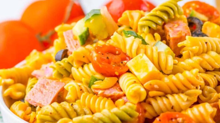 A close-up of a vibrant pasta salad with tri-color rotini, cherry tomatoes, cubed cheese, olives, and chunks of salami.