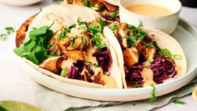 Two tacos filled with grilled chicken, shredded cabbage, fresh cilantro, and topped with creamy sauce, served on a plate with a small bowl of dipping sauce.