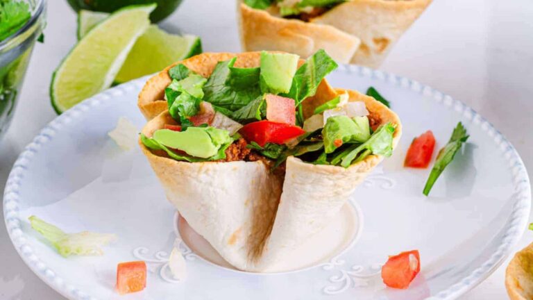 Mini taco bowl filled with lettuce, avocado, tomato, and meat, on a white plate. Lime slices and other bowls in the background.