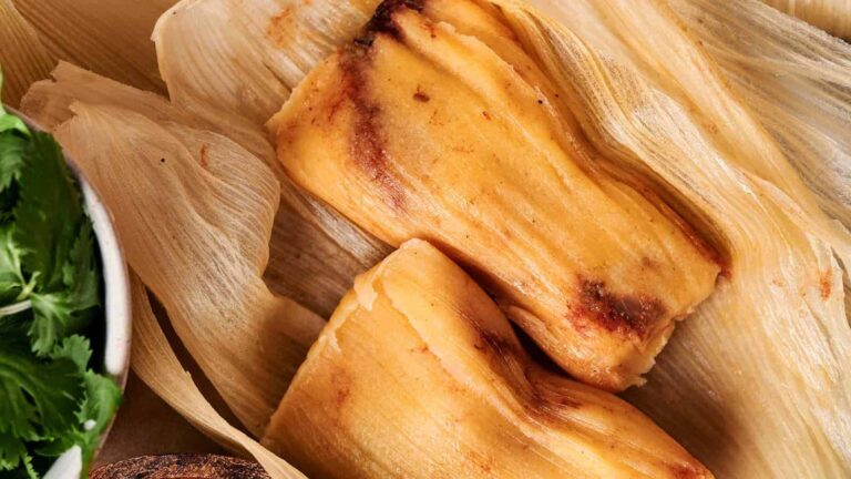 Two tamales wrapped in corn husks are placed on a surface. Fresh cilantro is visible on the side.
