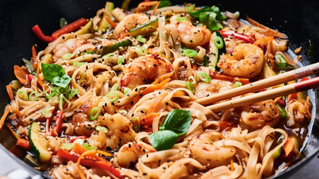 A close-up of stir-fried noodles with shrimp, vegetables, and herbs in a wok, garnished with sesame seeds and green onions. Chopsticks rest on the noodles.