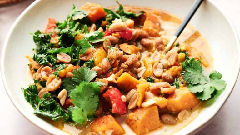 A bowl of curry with diced squash, leafy greens, red bell peppers, and peanuts, garnished with cilantro. A spoon rests in the curry.