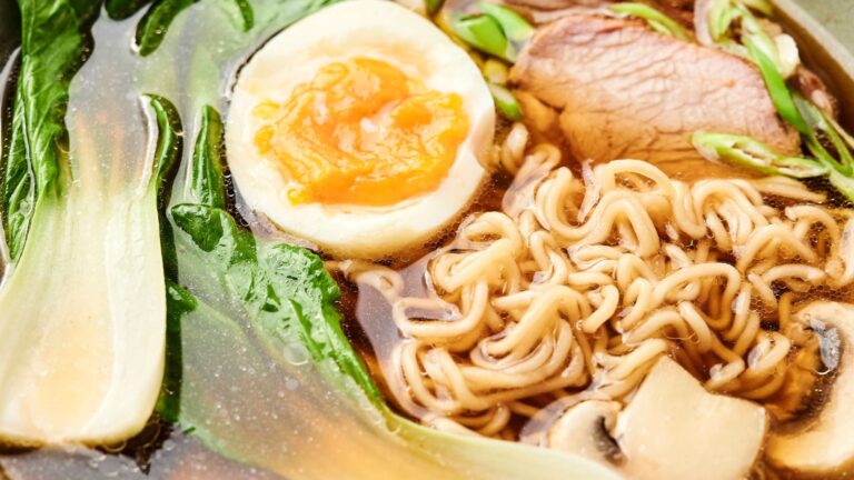 A bowl of ramen with noodles, sliced pork, a soft-boiled egg, bok choy, mushrooms, and green onions in broth.