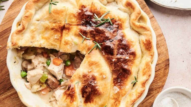 A baked chicken pot pie with a portion removed, revealing a filling of chicken, peas, mushrooms, and carrots. Garnished with rosemary on a wooden board.