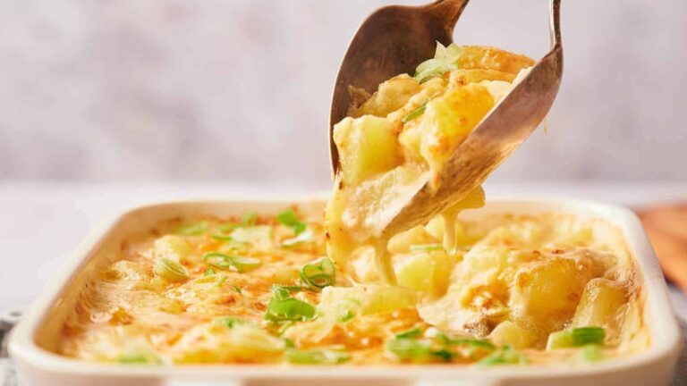 A spoon serves cheesy potato gratin from a baking dish, topped with melted cheese and chopped green onions.