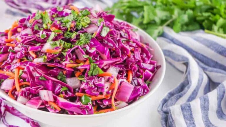 A bowl of vibrant purple cabbage salad with shredded carrots and chopped herbs, next to a striped cloth and fresh greens.