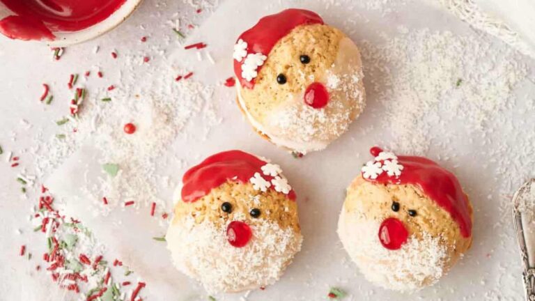 Three Santa-themed cookies decorated with red icing hats, white sprinkles, and candy faces on a white surface with scattered sprinkles.