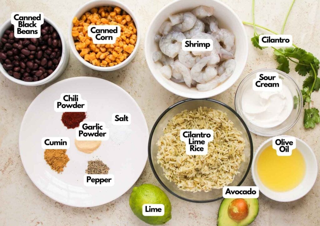 Various labeled ingredients for a Shrimp Burrito Bowl are arranged in bowls and on a counter, including shrimp, canned black beans, corn, cilantro lime rice, spices, cilantro, sour cream, olive oil, avocado, and lime.