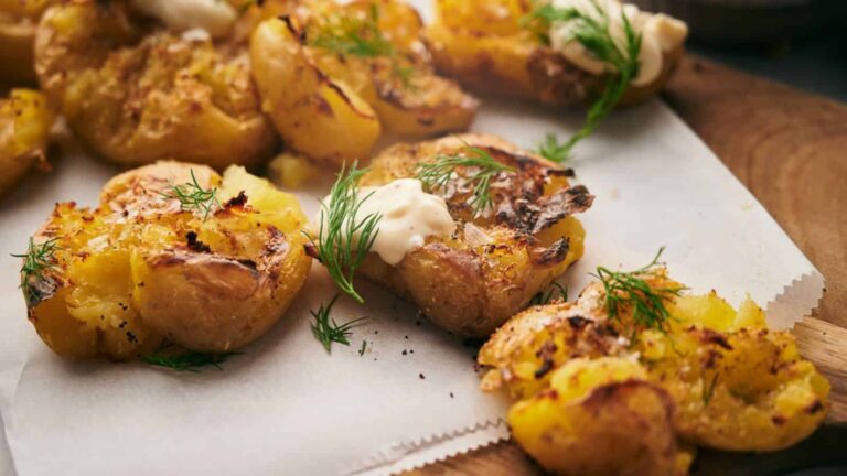 Close-up of smashed potatoes topped with a dollop of sour cream and garnished with fresh dill on a parchment-lined wooden board.