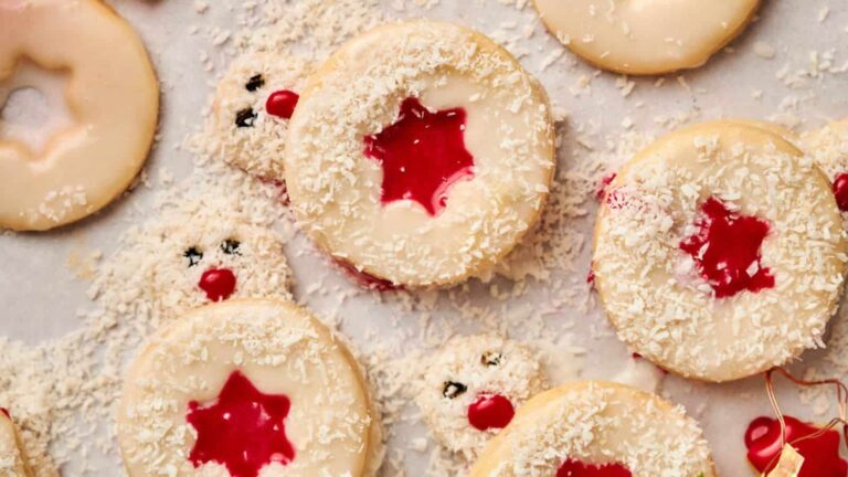 Cookies with red star-shaped centers and white frosting, sprinkled with coconut flakes, are arranged on a surface. Some have chocolate eyes, resembling festive reindeer.