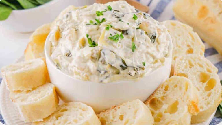 A white bowl of creamy spinach artichoke dip garnished with parsley, surrounded by slices of bread on a plate.