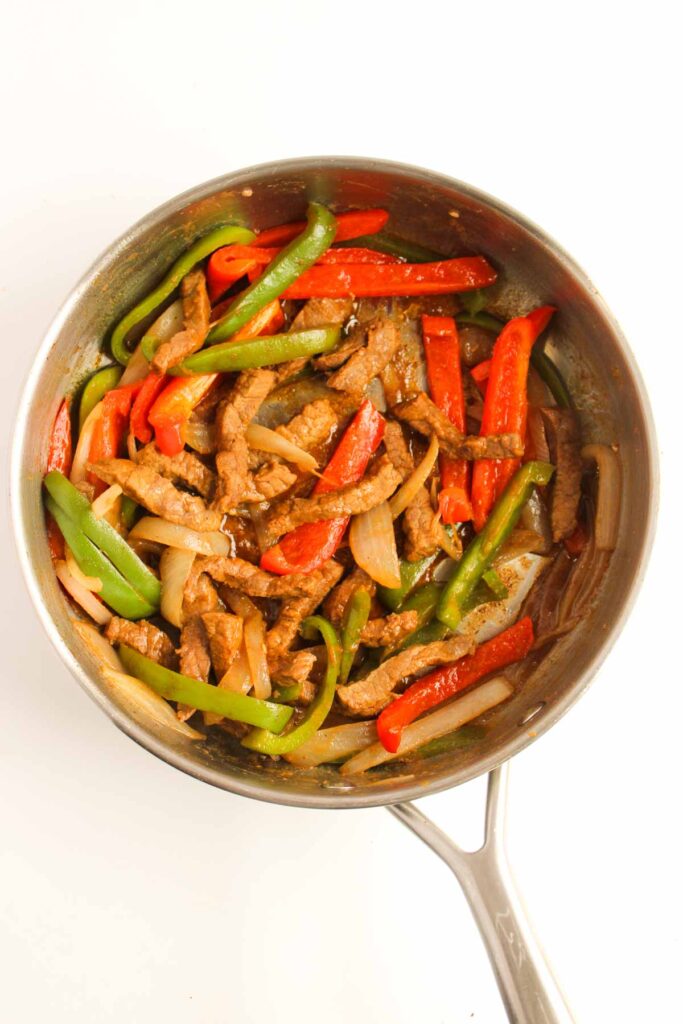 Steak fajita strips sizzle with red and green bell peppers and onions in a stainless steel pan.