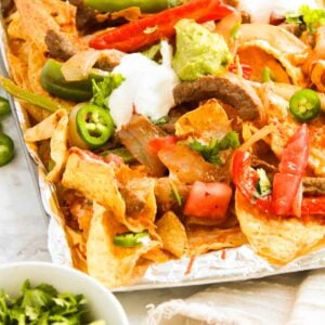 Close-up of steak fajita nachos topped with jalapeños, tomatoes, onions, guacamole, sour cream, and cilantro, all served on a foil-lined tray.